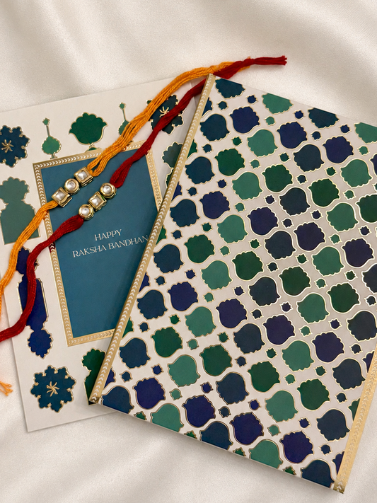 A festive Rakhi with a blue card and a patterned envelope on a white satin fabric background.