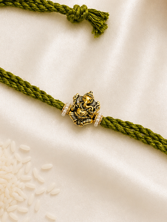 Antique gold-tone Ganesha Rakhi with pearl accents on an olive green braided thread, displayed on a silk background.