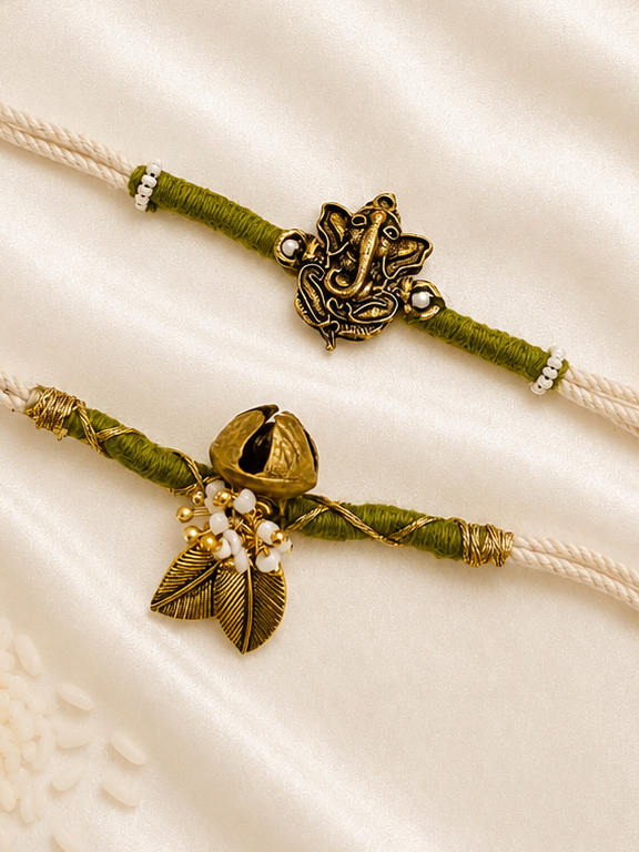 Bhai-Bhabhi Rakhi Set with an antique gold-tone Ganesha rakhi and Ghungroo bracelet with pearl and leaf accents on ivory and olive green threads, displayed on a silk background with kumkum and rice.
