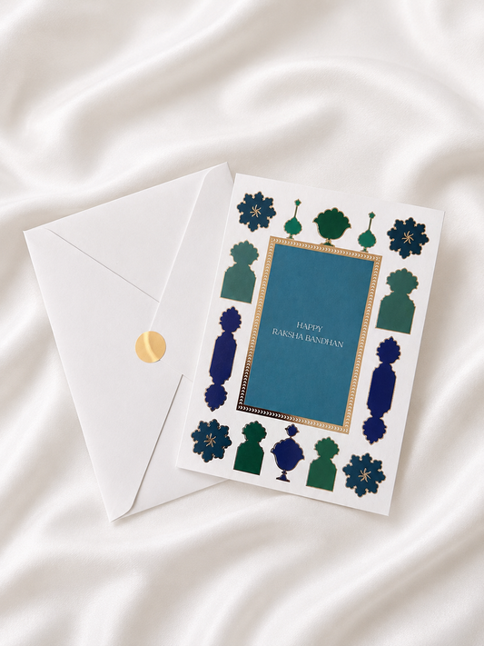 Premium Raksha Bandhan greeting card with blue and green gold foil accents and matching envelope, displayed on a white silk background.