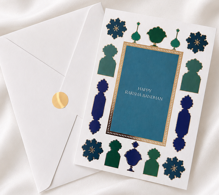 Premium Raksha Bandhan greeting card with blue and green gold foil accents and matching envelope, displayed on a white silk background.