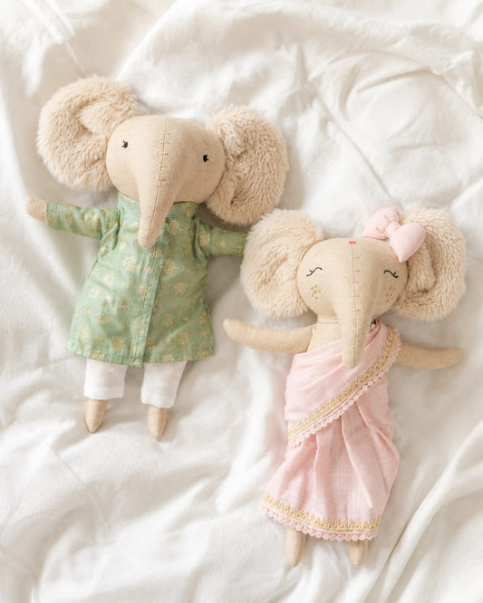 Two plush elephant dolls lying on white fabric. One doll wearing a green sherwani and one wearing a blush pink saree.