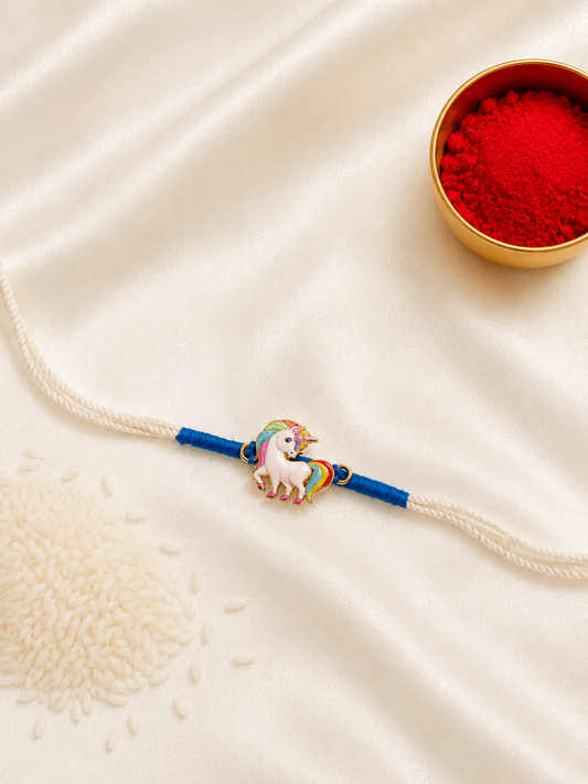 Kids Unicorn Rakhi with colorful enamel unicorn charm on a white thread with blue accents, displayed on a silk background with kumkum and rice.