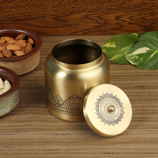 A brass-colored metal tea or spice container with a matching lid, both decorated with intricate mandala patterns, displayed on a wooden surface with decorative bowls of almonds and a green plant in the background.