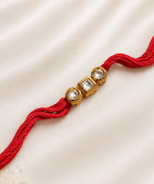 Three-Stone Kundan Rakhi with two round and one square bezel-set stones on a red thread, displayed on a silk background with rice.