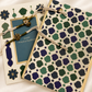 Two decorative greeting cards arranged on a cream satin fabric background. One card features a blue central panel with gold border and the text 'Happy Raksha Bandhan', while the other displays an intricate green, navy, and cream geometric pattern with gold borders and matching string ties.