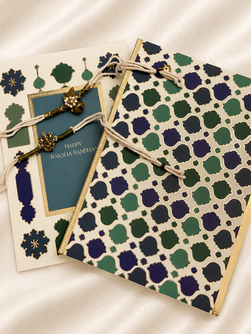 Two decorative greeting cards arranged on a cream satin fabric background. One card features a blue central panel with gold border and the text 'Happy Raksha Bandhan', while the other displays an intricate green, navy, and cream geometric pattern with gold borders and matching string ties.