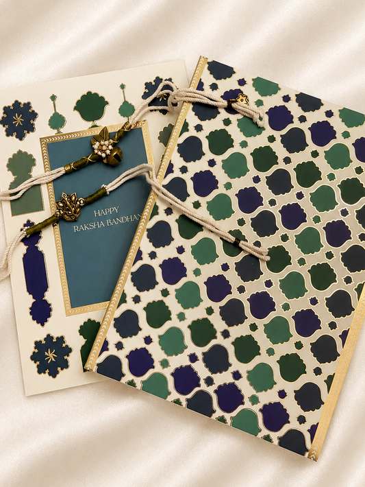 Two decorative greeting cards arranged on a cream satin fabric background. One card features a blue central panel with gold border and the text 'Happy Raksha Bandhan', while the other displays an intricate green, navy, and cream geometric pattern with gold borders and matching string ties.
