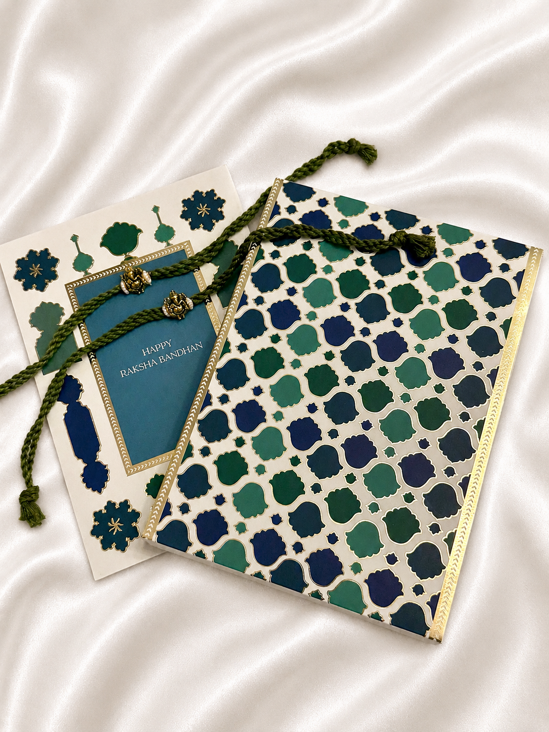 A Raksha Bandhan greeting card and matching envelope set arranged on a white satin fabric background. The envelope features a cream base with a repeating geometric trellis pattern in navy, teal, and green, finished with a gold border.