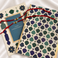 Two decorative greeting cards arranged on a white satin fabric background. One card has a blue center panel with gold text reading 'HAPPY RAKSHA BANDHAN,' and the other has an allover geometric pattern in cream, teal, green, and navy with gold trim.