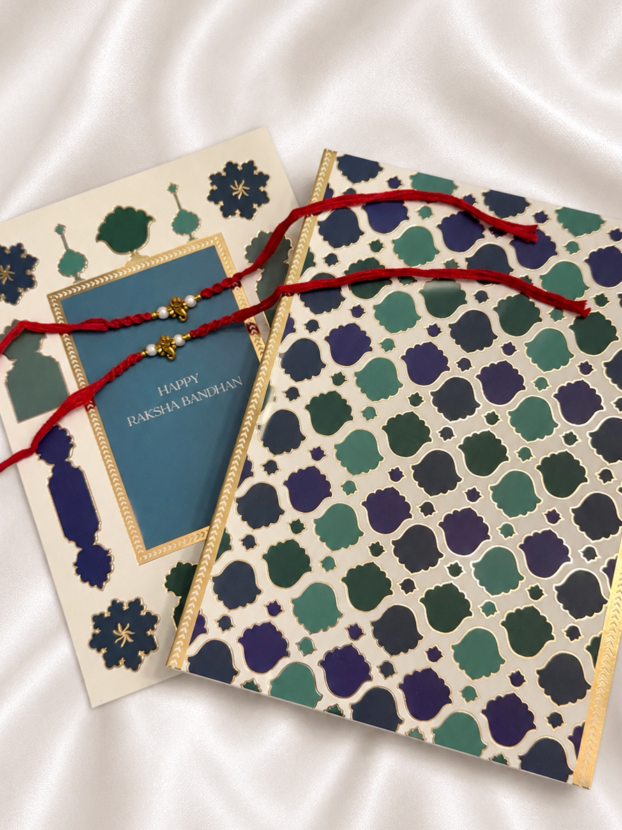 Two decorative greeting cards arranged on a white satin fabric background. One card has a blue center panel with gold text reading 'HAPPY RAKSHA BANDHAN,' and the other has an allover geometric pattern in cream, teal, green, and navy with gold trim.