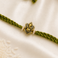 Antique gold-tone Ganesha Rakhi with pearl accents on an olive green braided thread, displayed on a silk background.
