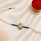Kids Ganesha Rakhi with colorful enamel Lord Ganesha charm on a white thread with teal accents, displayed on a silk background with kumkum and rice.