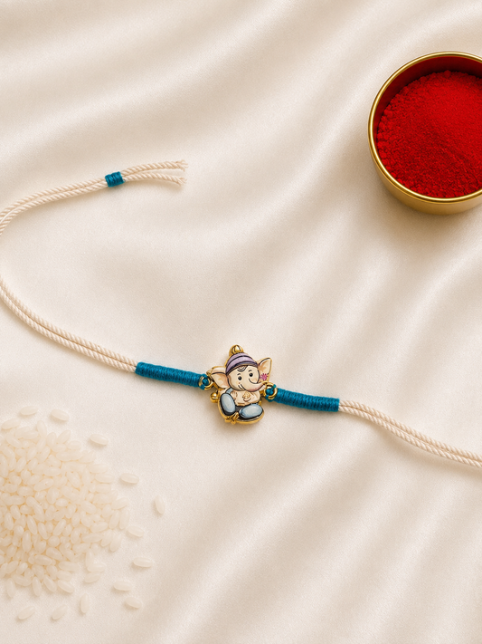 Kids Ganesha Rakhi with colorful enamel Lord Ganesha charm on a white thread with teal accents, displayed on a silk background with kumkum and rice.