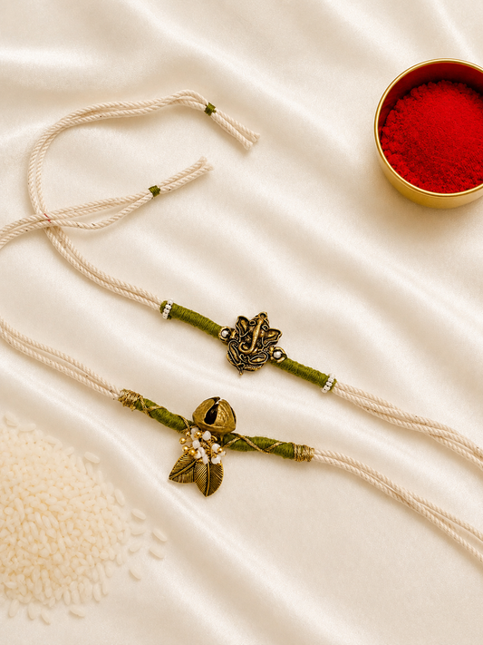 Bhai-Bhabhi Rakhi Set with an antique gold-tone Ganesha rakhi and Ghungroo bracelet with pearl and leaf accents on ivory and olive green threads, displayed on a silk background with kumkum and rice.
