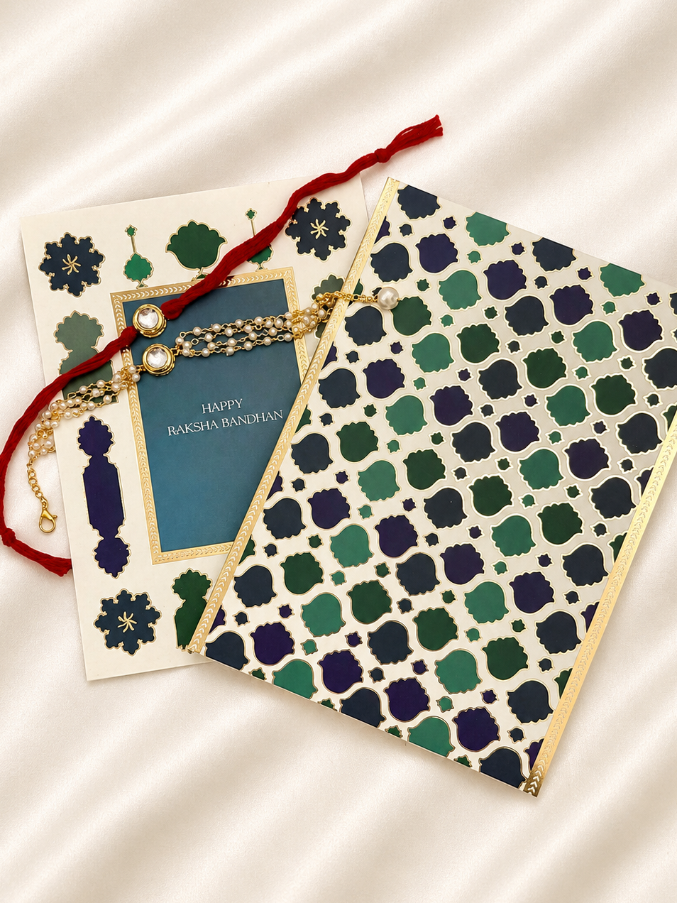 A festive Raksha Bandhan greeting card and envelope set with a red and gold Rakhi laid across them, styled on a cream satin fabric background.