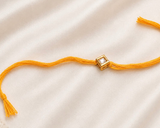 Single Square Kundan Rakhi with a bezel-set square stone on a yellow thread, displayed on a silk background.