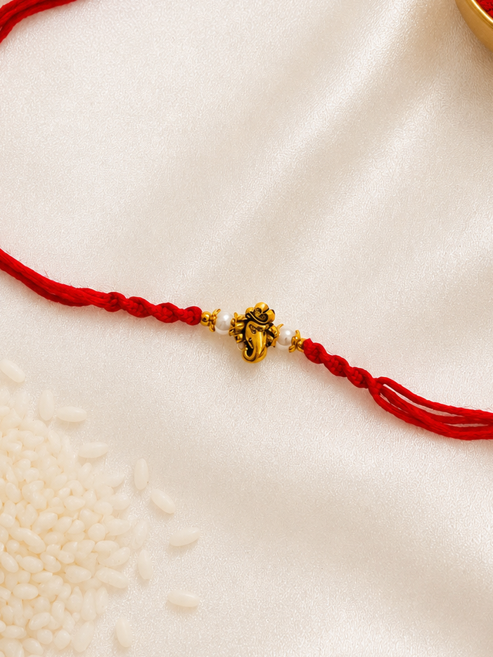 Gold-tone Ganesha Rakhi on a red thread, displayed on a silk background.