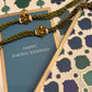 A close-up of a Raksha Bandhan gift set featuring a blue card with gold foil borders and 'HAPPY RAKSHA BANDHAN' text, wrapped with an olive green braided cord and gold-plated decorative beads, alongside patterned wrapping paper.