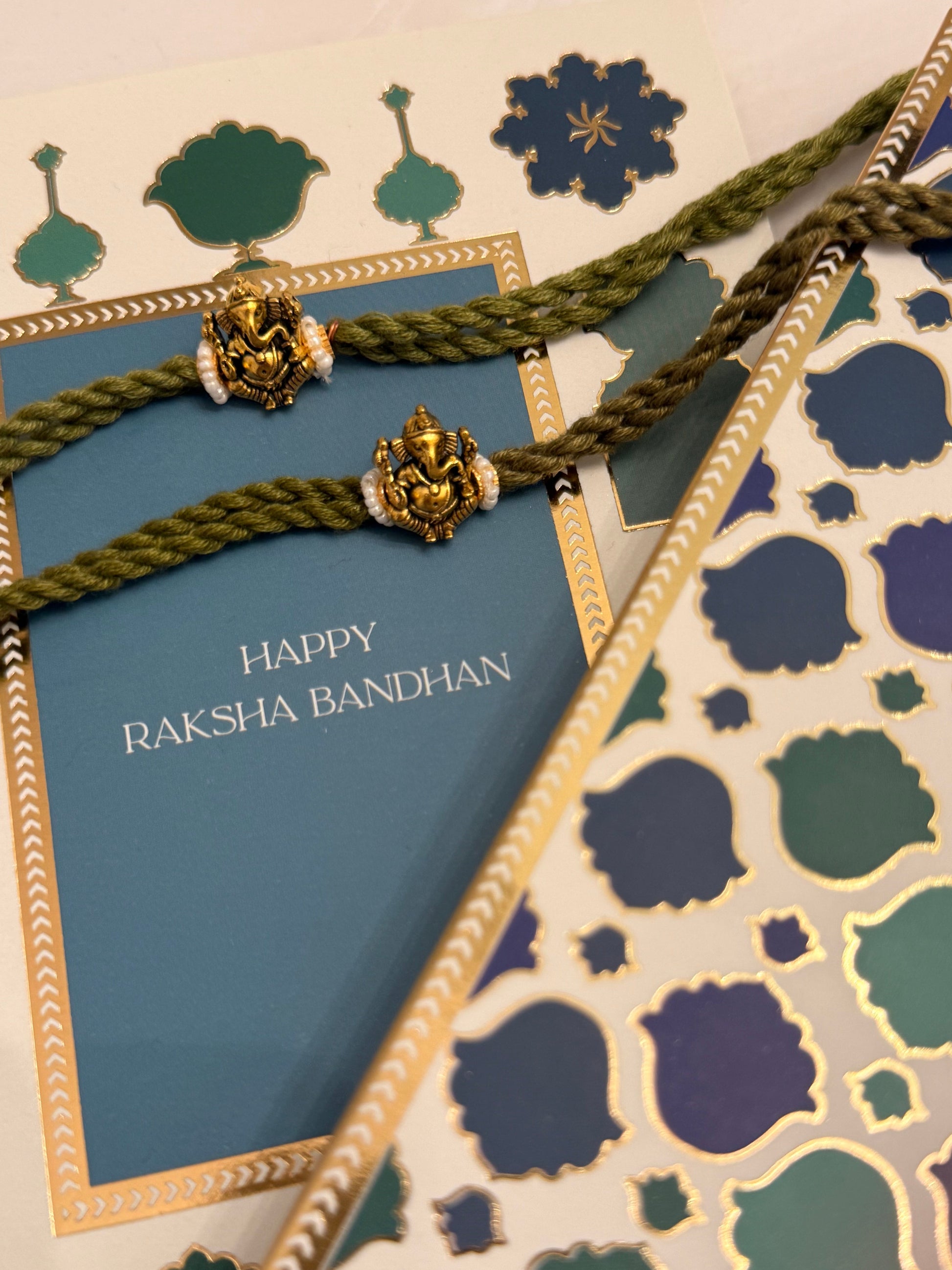 A close-up of a Raksha Bandhan gift set featuring a blue card with gold foil borders and 'HAPPY RAKSHA BANDHAN' text, wrapped with an olive green braided cord and gold-plated decorative beads, alongside patterned wrapping paper.