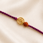 Gold-tone Om and Rudraksha Rakhi on a purple braided thread, displayed on a silk background.