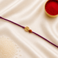 Gold-tone Om and Rudraksha Rakhi on a purple braided thread, displayed on a silk background with kumkum and rice.