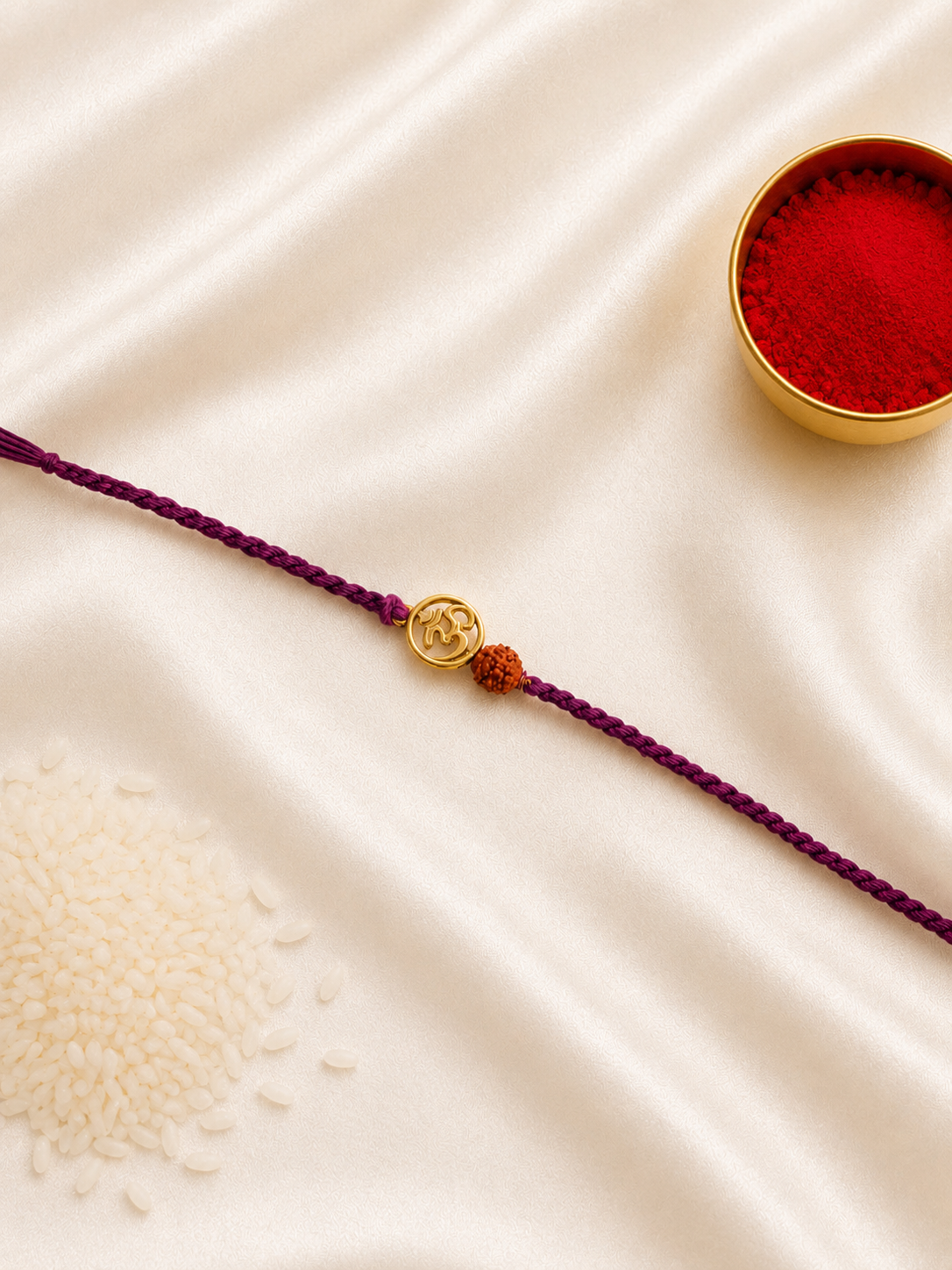Gold-tone Om and Rudraksha Rakhi on a purple braided thread, displayed on a silk background with kumkum and rice.
