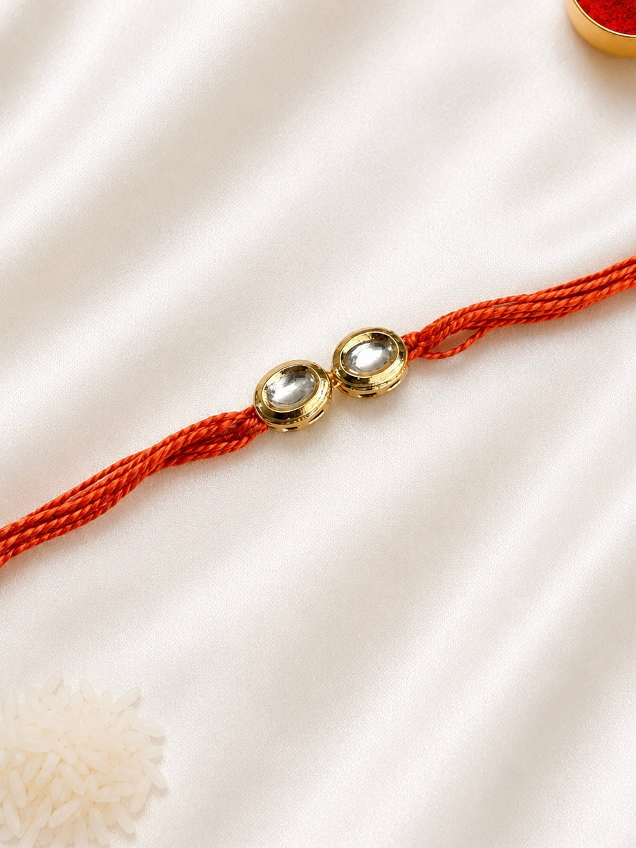 Twin Round Kundan Rakhi with two round bezel-set stones on a rust-orange thread, displayed on a silk background with kumkum and rice.
