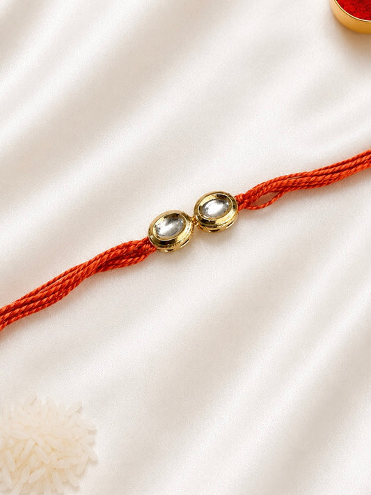 Twin Round Kundan Rakhi with two round bezel-set stones on a rust-orange thread, displayed on a silk background with kumkum and rice.