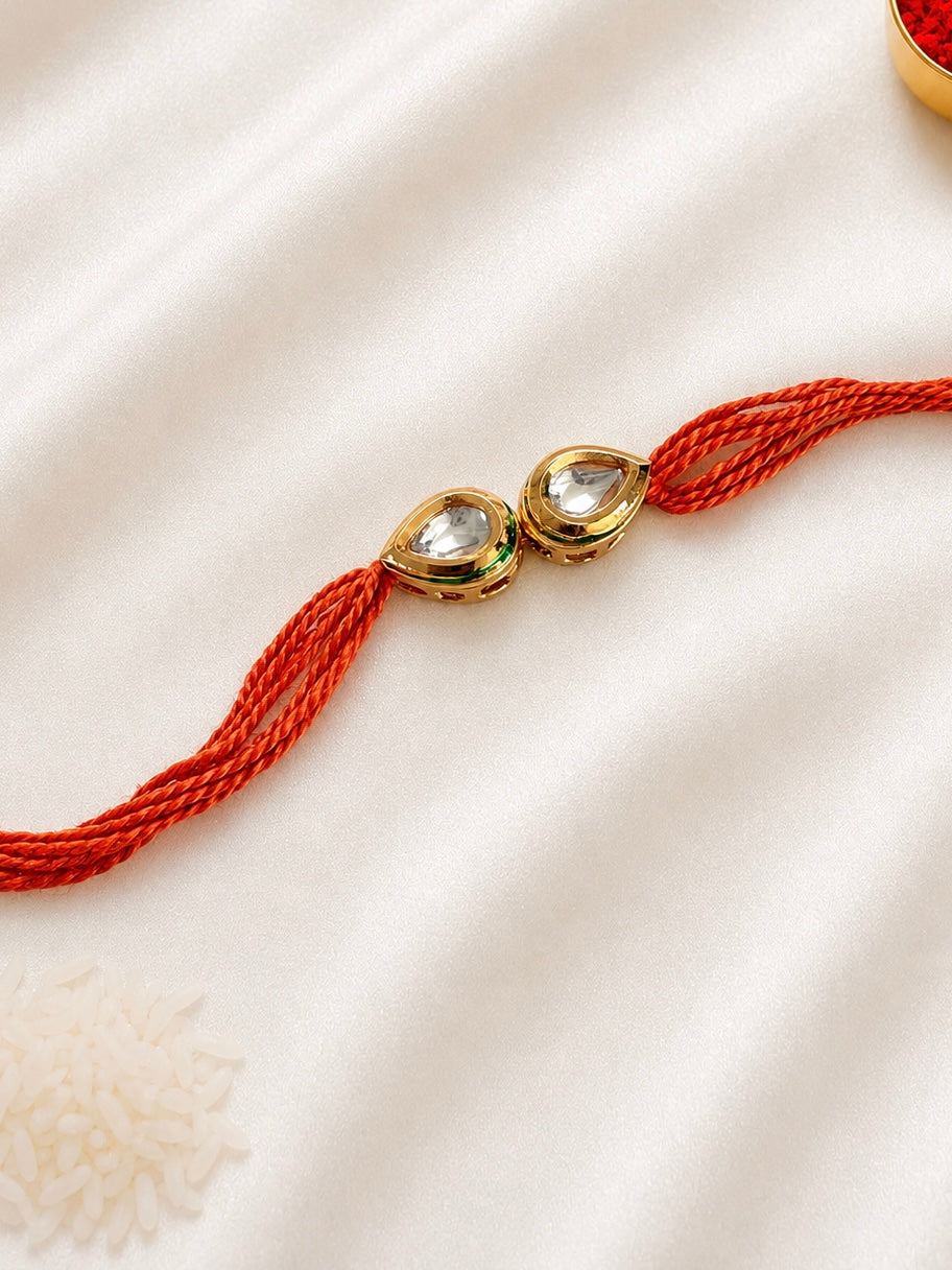 Twin Pear Kundan Rakhi with two pear-shaped bezel-set stones on a rust-orange thread, displayed on a silk background with kumkum and rice.