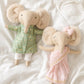 Two plush elephant dolls lying on white fabric. One doll wearing a green sherwani and one wearing a blush pink saree.