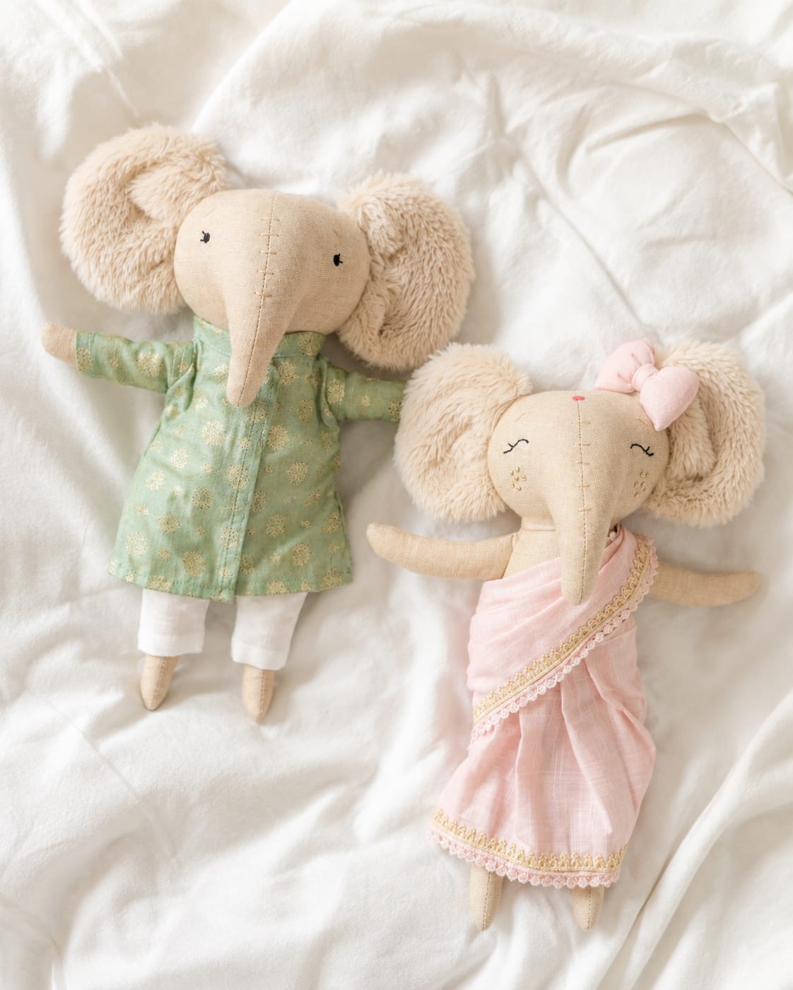 Two plush elephant dolls lying on white fabric. One doll wearing a green sherwani and one wearing a blush pink saree.