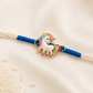 Kids Unicorn Rakhi with colorful enamel unicorn charm on a white thread with blue accents, displayed on a silk background.