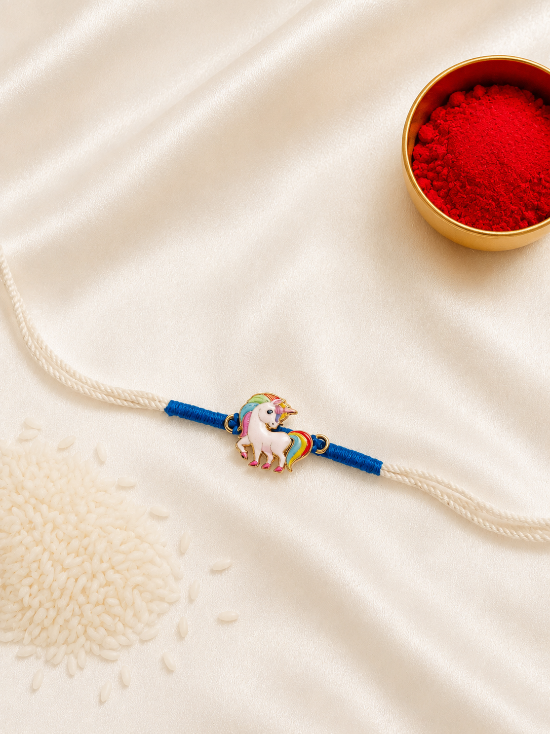 Kids Unicorn Rakhi with colorful enamel unicorn charm on a white thread with blue accents, displayed on a silk background with kumkum and rice.