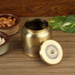 A brass-colored metal tea or spice container with a matching lid, both decorated with intricate mandala patterns, displayed on a wooden surface with decorative bowls of almonds and a green plant in the background.